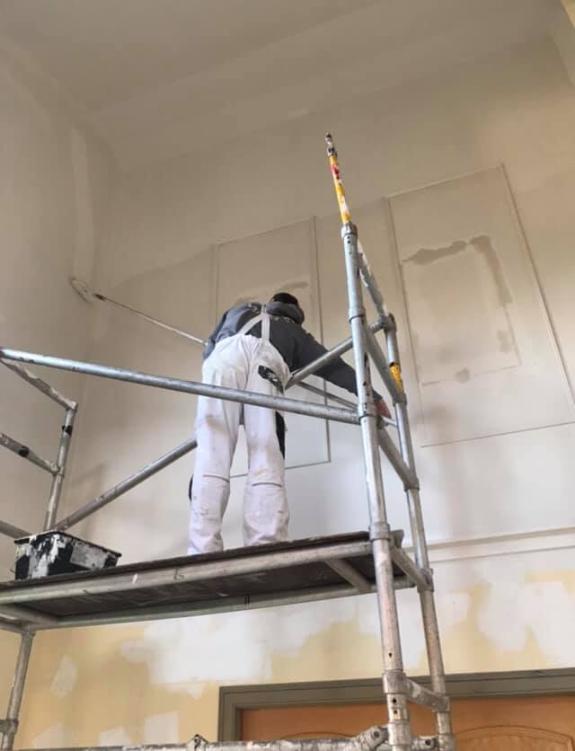 A man in paint overalls, standing on scaffolding inside a building to paint the walls